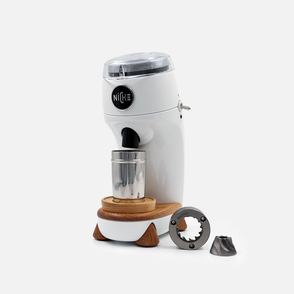 The Niche Zero single-dose conical burr grinder in its walnut-wood and matte-metal finish, shown from a three-quarter front angle with its catch cup in place beneath the exit chute.